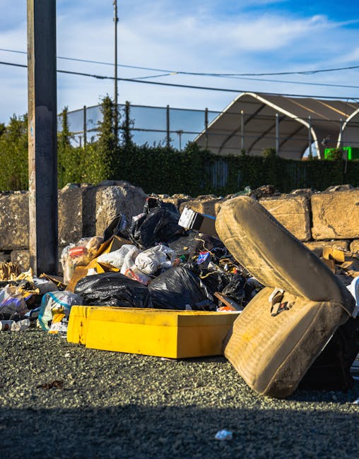 A waste collection worker, dressed in a red and yellow high-visibility vest, is seen operating a large red rubbish truck on a roadside, standing next to the open rear compartment where waste materials