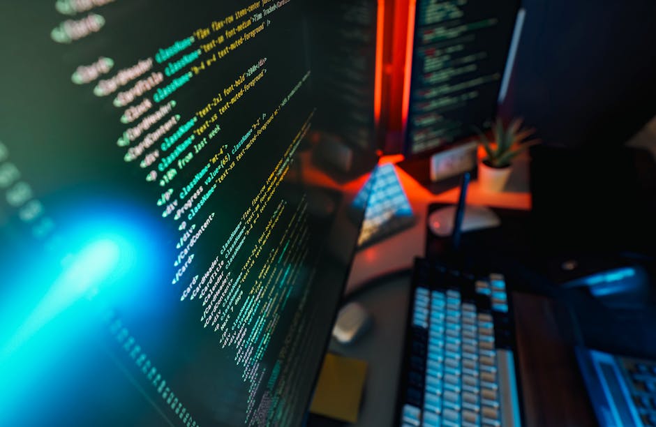 A person seated at a desk, viewed from behind and to the left, working on a laptop with a code editor visible on the screen displaying multicolored lines of programming code. The individual is wearing