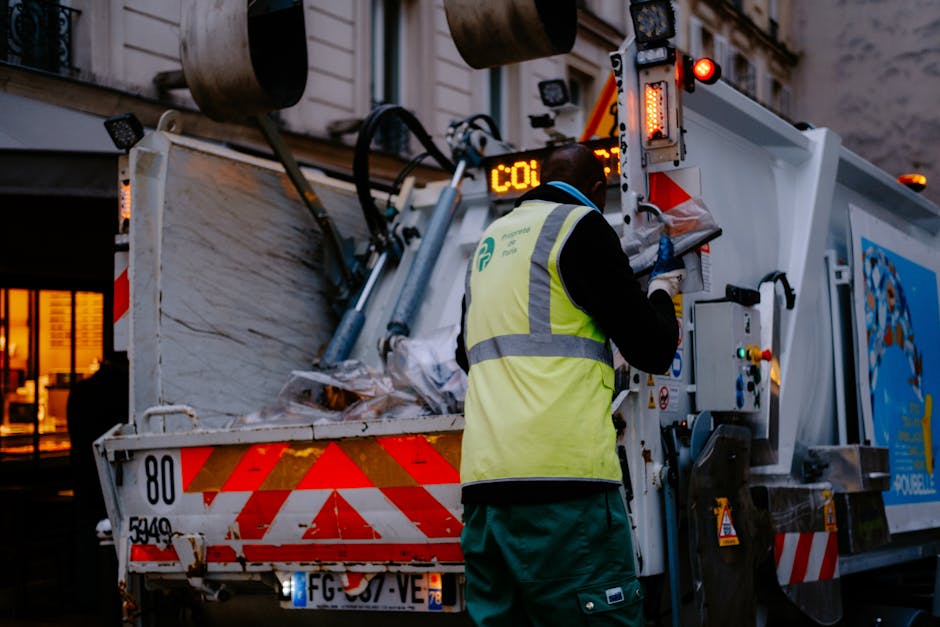 A large, yellow excavator equipped with a thick, textured metal bucket is lifting a pile of cut, snow-like material, possibly waste or snow, into an orange dump truck parked outdoors. The material app